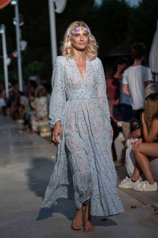 A woman wearing a light, floral-patterned bohemian gown walking barefoot at La Galeria Elefante Ibiza - Atzaro fashion show Ibiza surrounded by an engaged crowd.