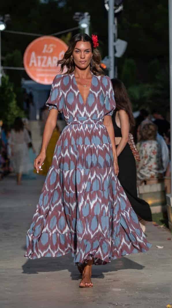A woman walks the runway in a colorful, flowing maxi dress with blue, purple, and white patterns for La Galeria Elefante Ibiza - Atzaro fashion show Ibiza