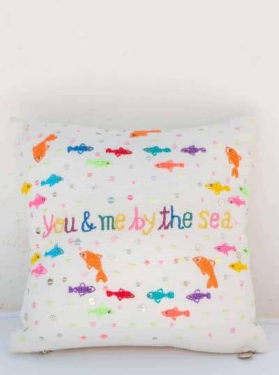 small square cushion with colourful embroidered fish on it that says you and me by the sea