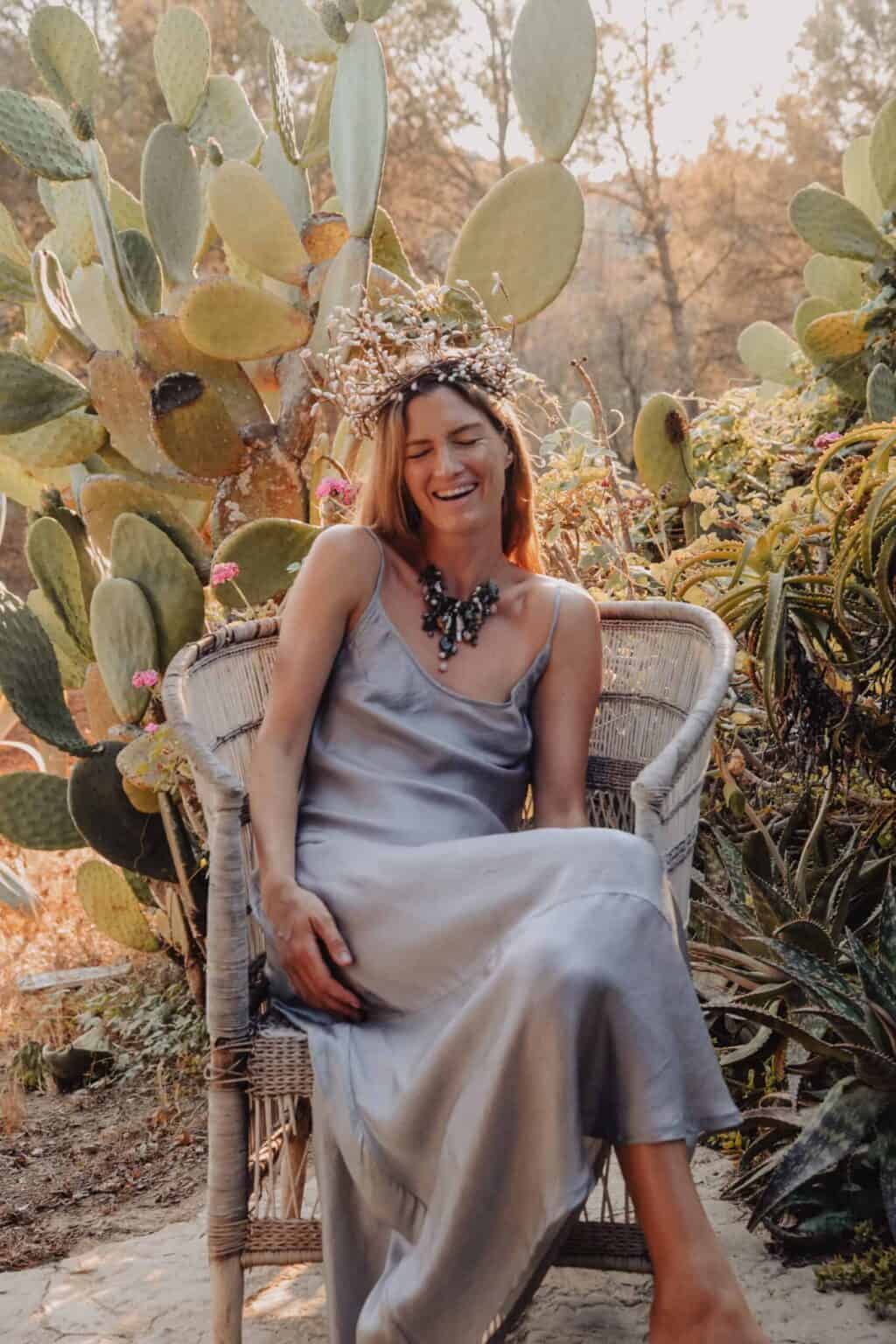 Beautiful woman sitting in vintage chair among cactus plants in a lush garden wearing a long grey silk dress.