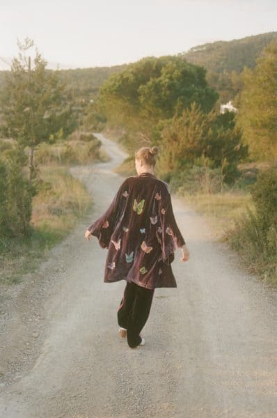 A woman walking down a countryside dirt road surrounded by trees and hills during sunset, wearing a butterfly-printed kimono for a serene outdoor experience.