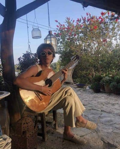 A person sitting on a wooden chair playing guitar in a serene garden environment with hanging lanterns and flowering trees.