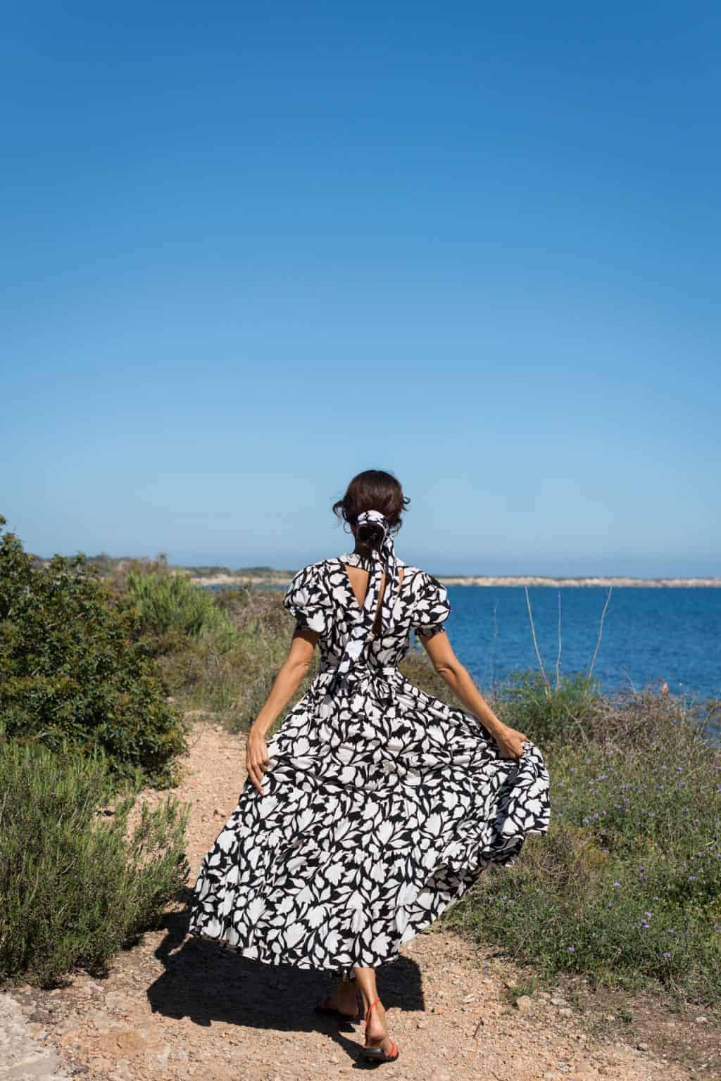 Beautiful woman in black and white floral dress walking on coastal trail with sea view for La Galeria Elefante, stylish beach fashion and scenic landscape.