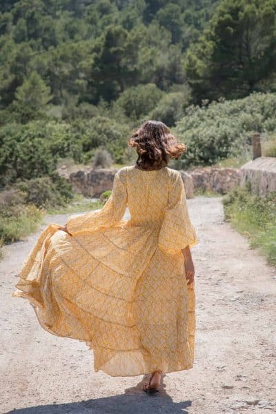 Enchanting woman with Dark hair walking on nature trail in a flowing yellow dress, showcasing the natural beauty and artistic design of La Galeria Elefante. Perfect for bohemian fashion and nature-inspired clothing.