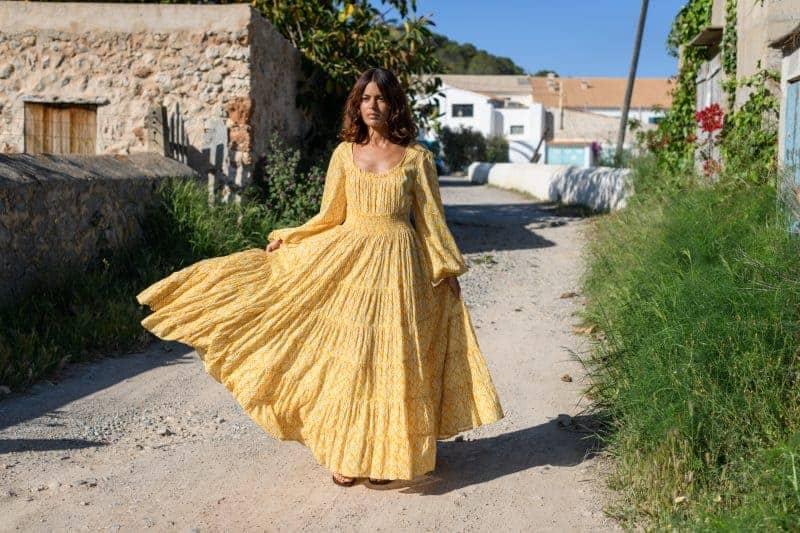 Colorful yellow bohemian dress on a woman walking on a rustic village street with white houses and greenery, showcasing vibrant fashion and artistic style. La Galeria Elefante ibiza John Lewis x La Galeria Elefante Capsule Collection