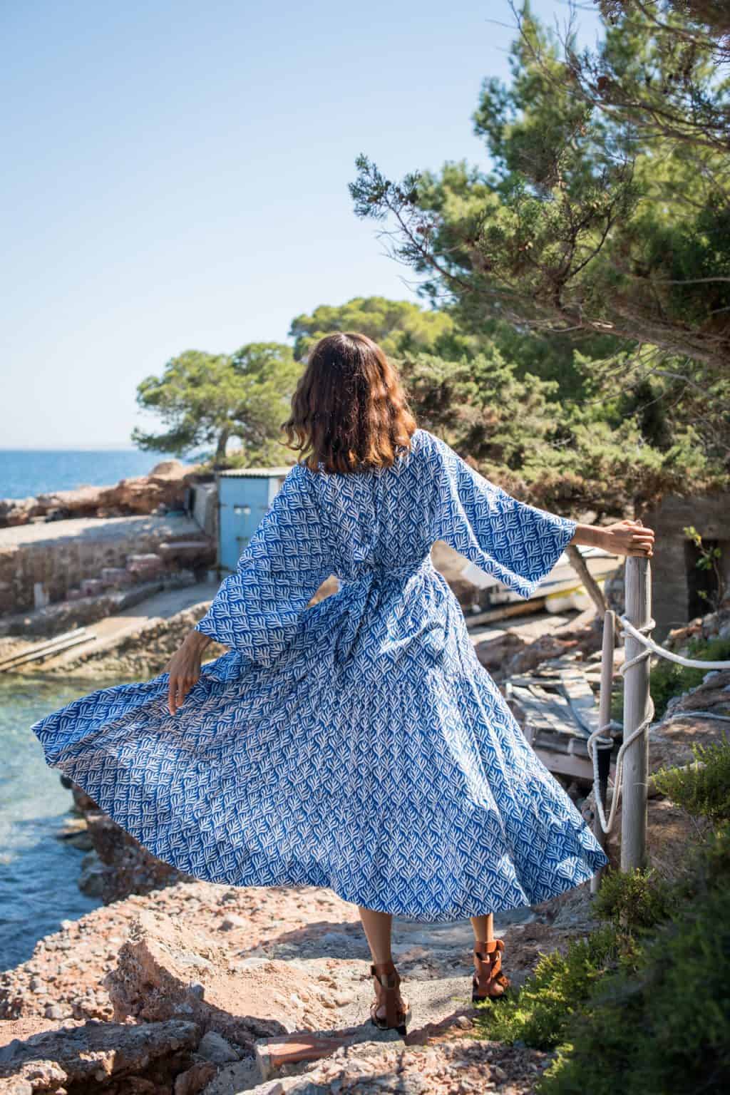 A woman in a blue and white patterned dress walking along a rocky coastline with lush greenery and trees in the background.