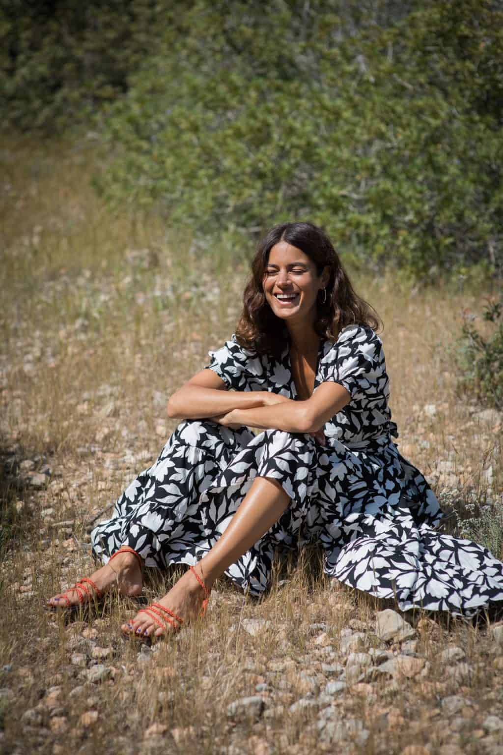 Vibrant woman in a black and white floral dress sitting on dry grass in nature, sunny outdoor scenery, joyful mood, and lifestyle photography for La Galeria Elefante.