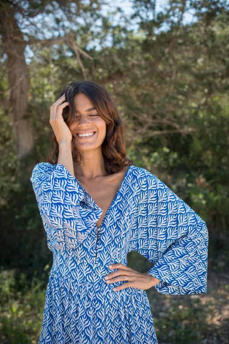 Playful woman smiling in flowy blue patterned dress outdoors at La Galeria Elefante, surrounded by greenery, perfect for summer fashion and nature lovers.