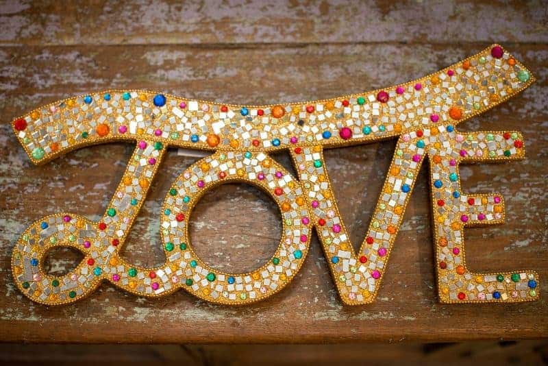 Colorful mosaic "LOVE" sign with rhinestones and mirror pieces, from La Galeria Elefante decor, arts, craft, or boutique event displays, on rustic wooden background.
