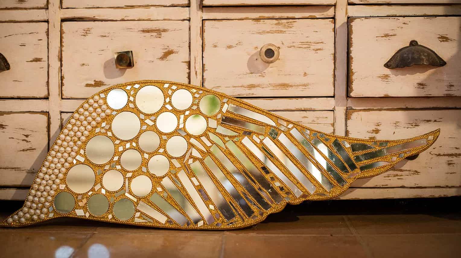 Colorful decorative mirror shaped like a wing, showcasing intricate mirror and pearl details, placed in front of vintage white wooden drawers.