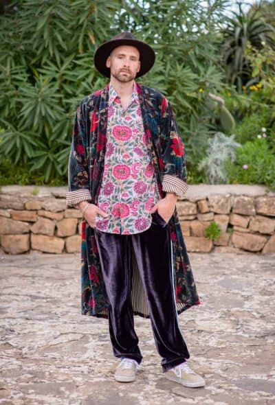 Model wearing purple and blue velvet trousers a black printed velvet dressing gown smoking jacket with a cotton floral block print shirt and a brown hat