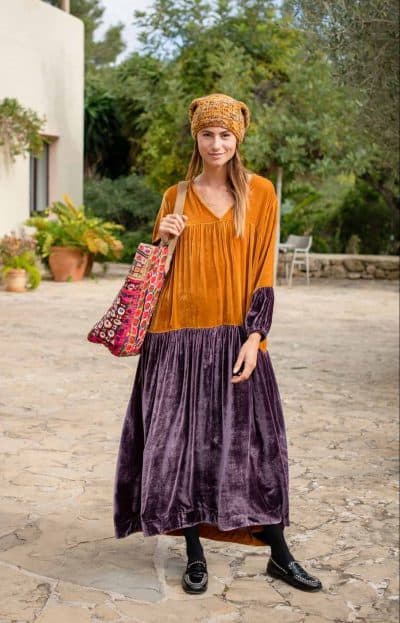 Model wears a floor length mustard and plum velvet dress with long sleeves and a yellow woolly beanie