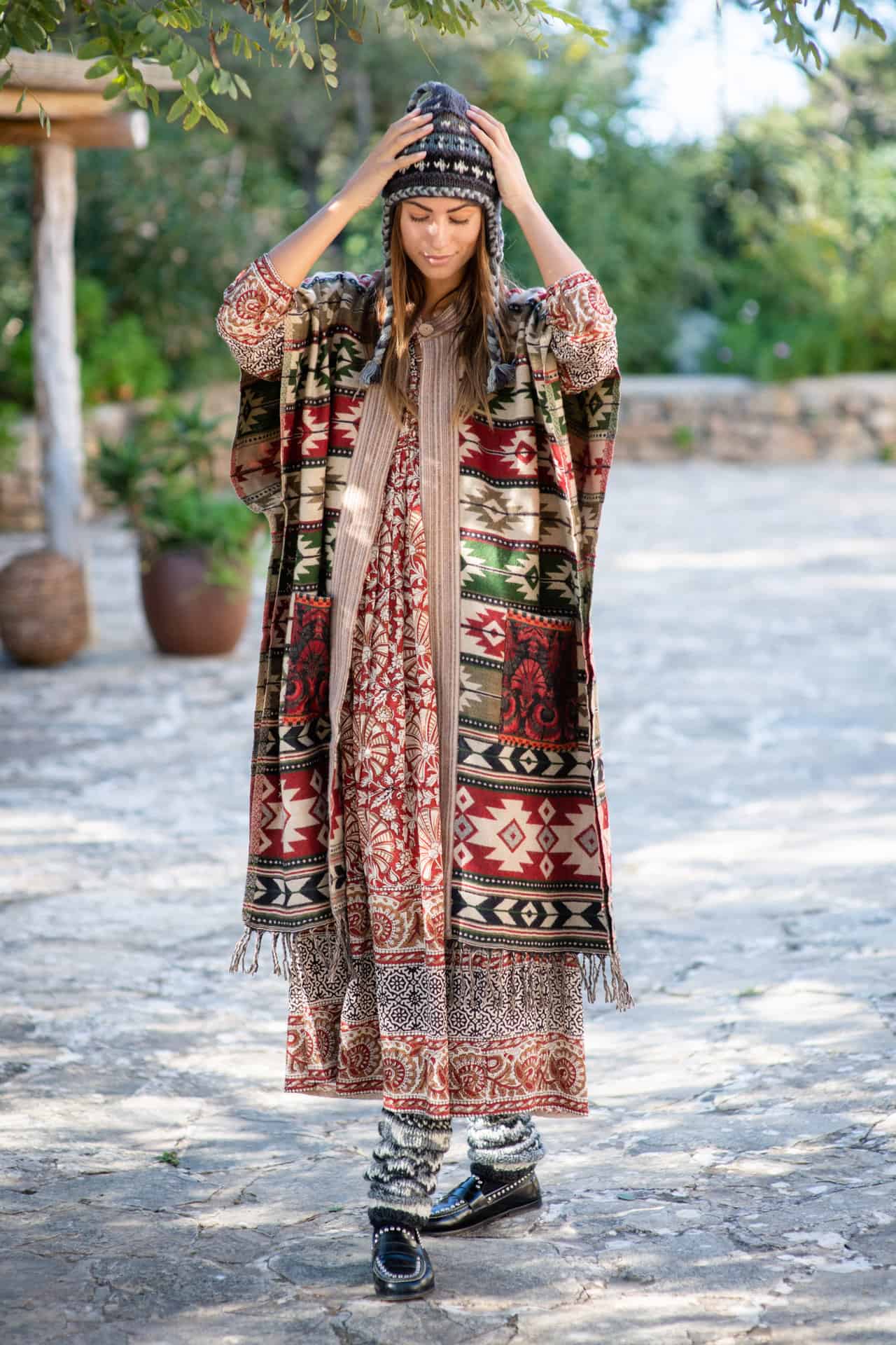 Model wears a block print floor length dress a poncho a woolly hat and woolly leg warmers in a garden