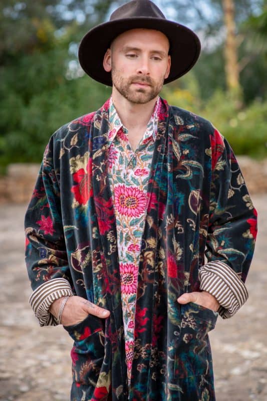 Model wears a black velvet printed dressing gown smoking jacket with a cotton floral block print shirt and a brown hat close up