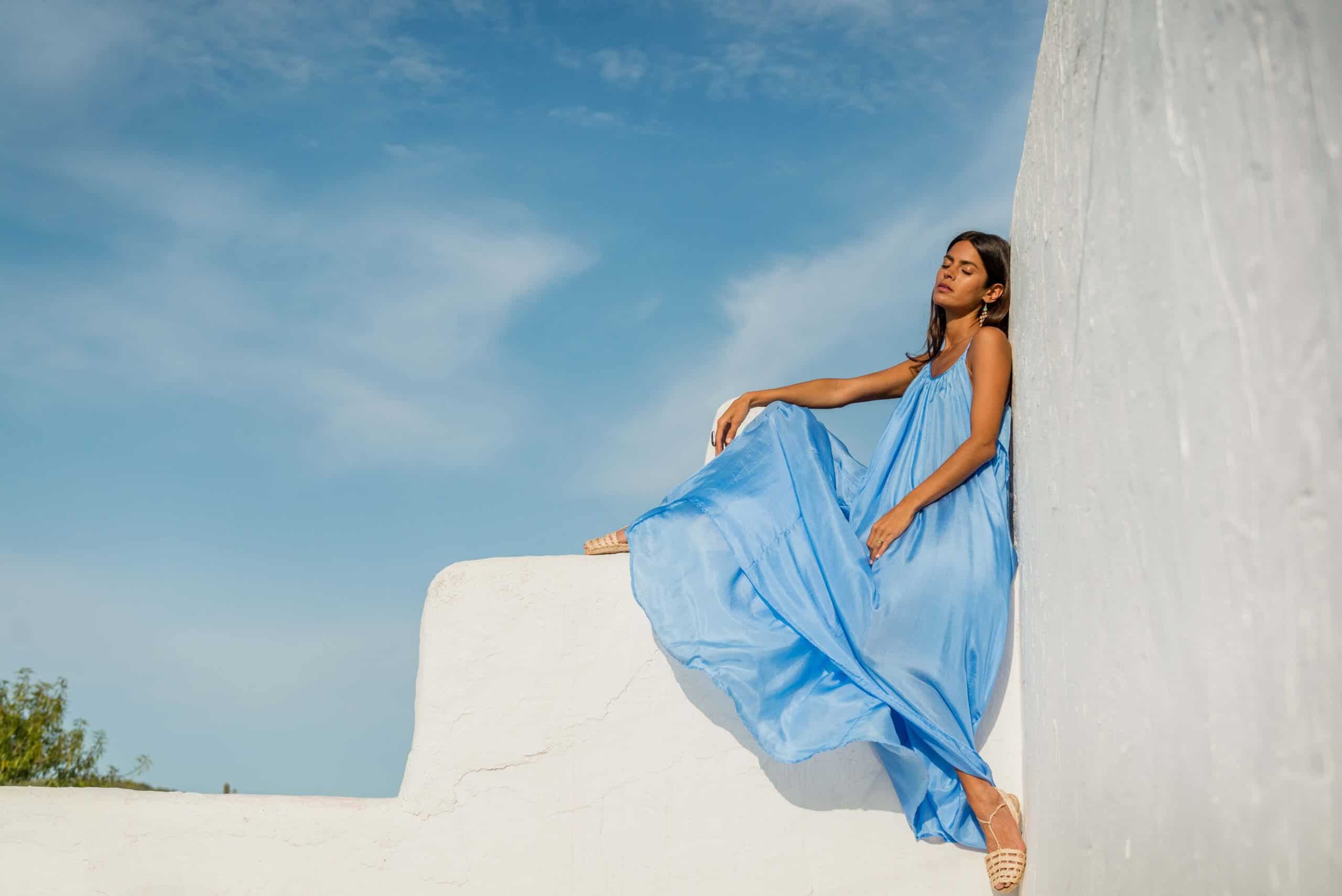 Model wearing a floor length blue silk dress sitting on a wall