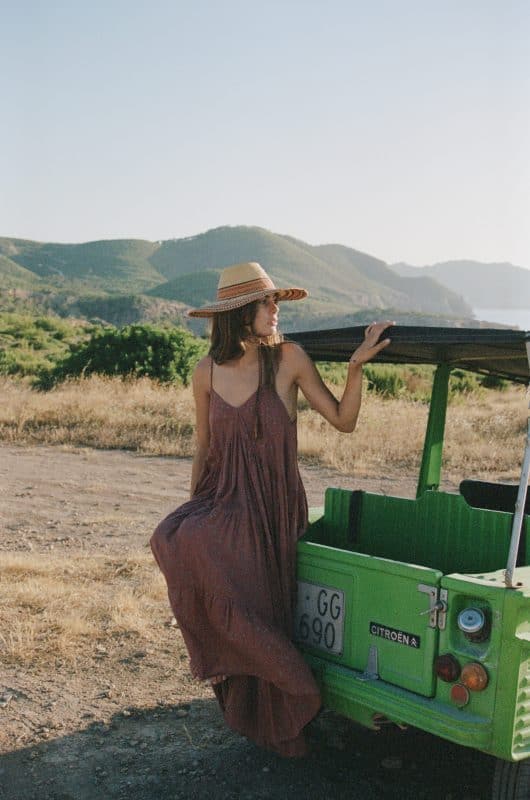 Model sitting on a green car wearing a strappy brown starry block print floor length cotton dress with a straw hat in a field