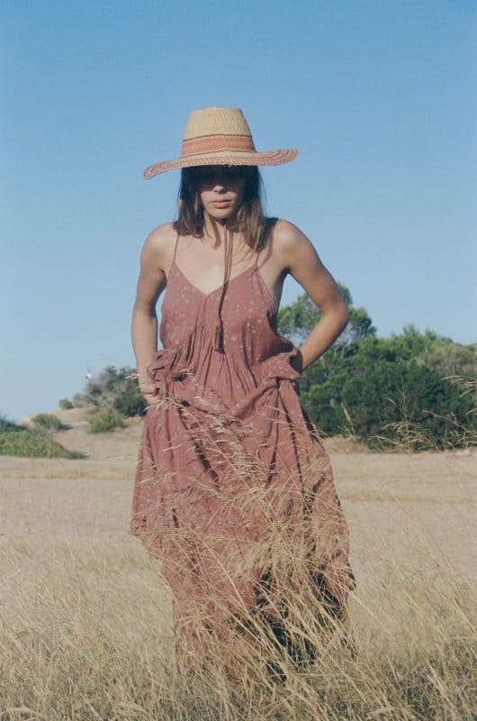 Model in a field wearing a strappy brown cotton block print starry dress with a straw hat in a field