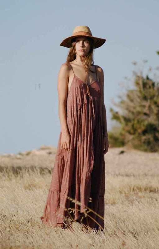 Model wearing a strappy floor length starry hand block print brown cotton dress with a hat in a field