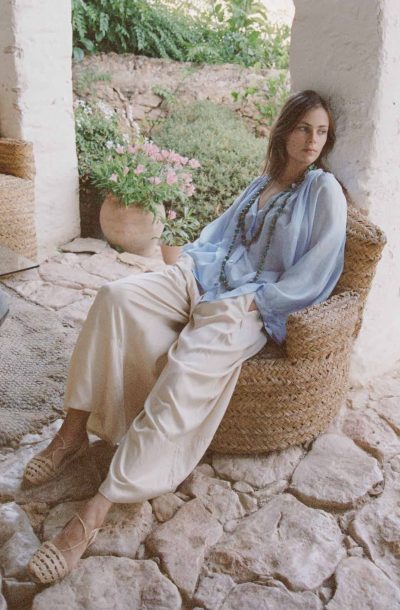 Model sitting down wearing cream modal wide leg trousers with a silk blouse in sky blue with a turquoise necklace