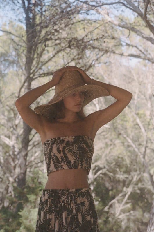 Model wearing skirt and boob tube set in green and cream leaf print with a straw hat in a forest