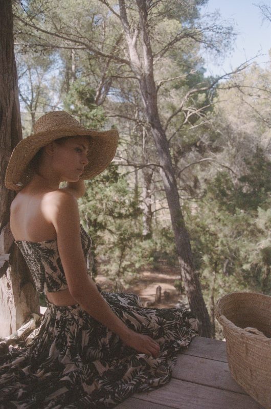 Model wearing a boob tube and skirt set in a green and cream leaf print with a straw hat sitting down profile