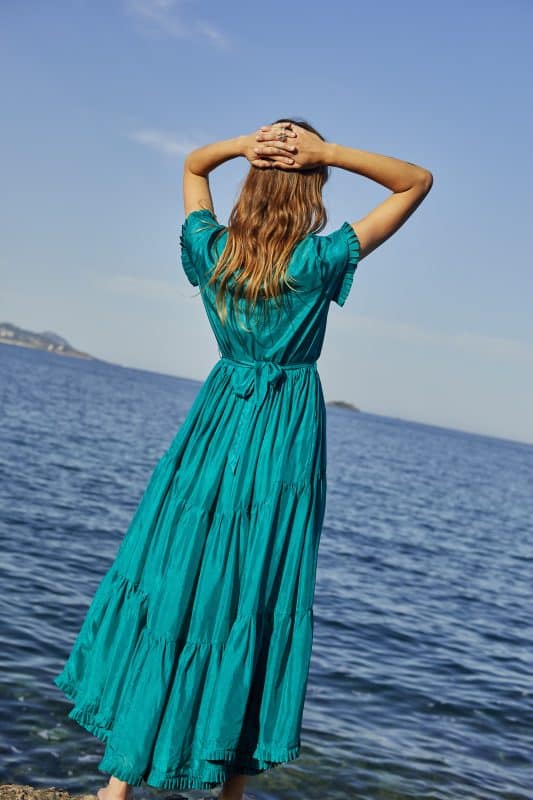 Back view of a model wearing a silk bottle green floor length tiered dress with short sleeves with a tie by the sea