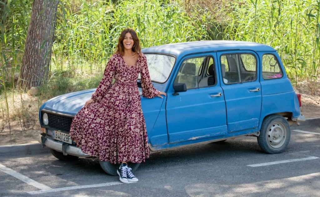La Galeria Elefante Ibiza Woman in floral dress leaning on vintage blue car in nature setting.