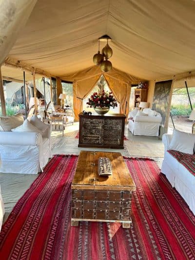 Luxurious vintage-style tent interior, with elegant white sofas, rustic wooden coffee table, vibrant red rugs, and floral centerpiece, perfect for upscale outdoor events and glamping.