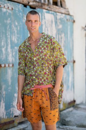 A vibrant image of a man wearing Jerome Shorts in Kantha, featuring colorful, patterned shorts and a matching shirt, standing outdoors against a rustic, weathered wall.