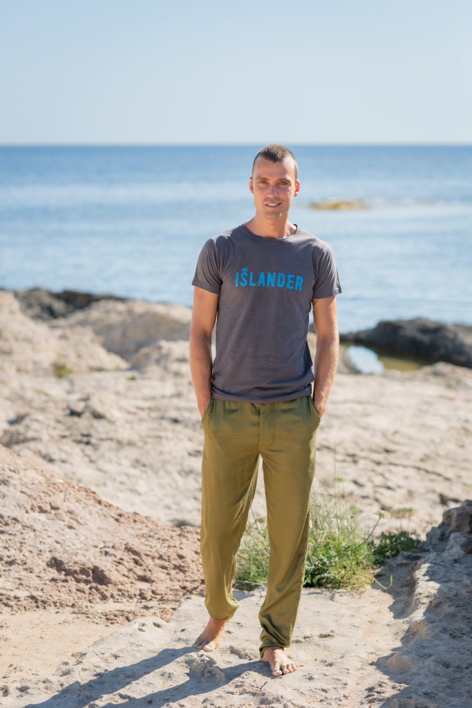 A person standing on a sandy beach near the ocean, wearing casual olive green trousers and a gray T-shirt with "ÍSLANDER" printed on it, under a clear blue sky.