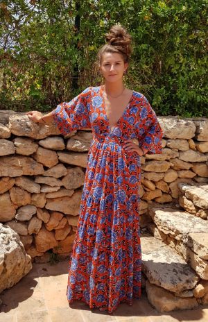 Elegant woman in bohemian maxi dress posing outdoors at La Galeria Elefante, Catalonia, Spain, surrounded by rustic stone wall and lush greenery.