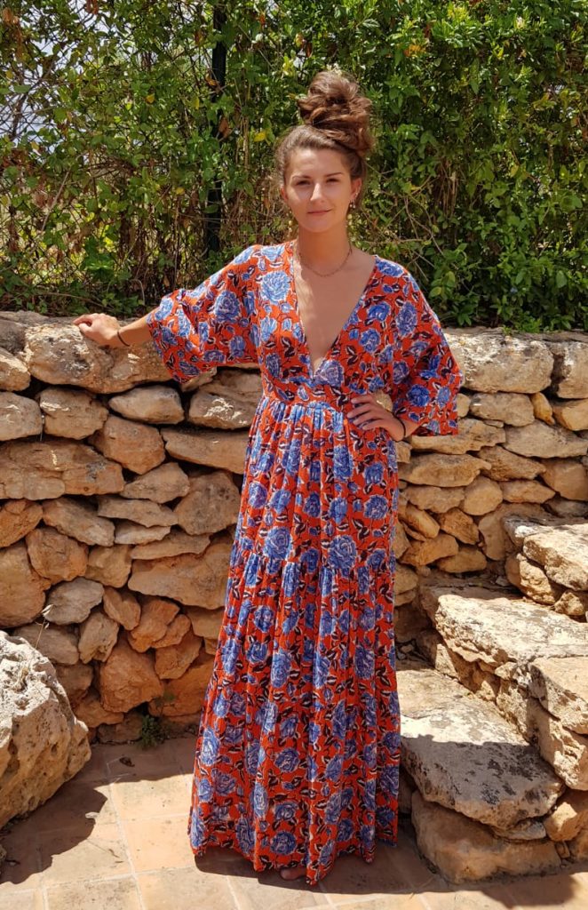 Elegant woman in bohemian maxi dress posing outdoors at La Galeria Elefante, Catalonia, Spain, surrounded by rustic stone wall and lush greenery.