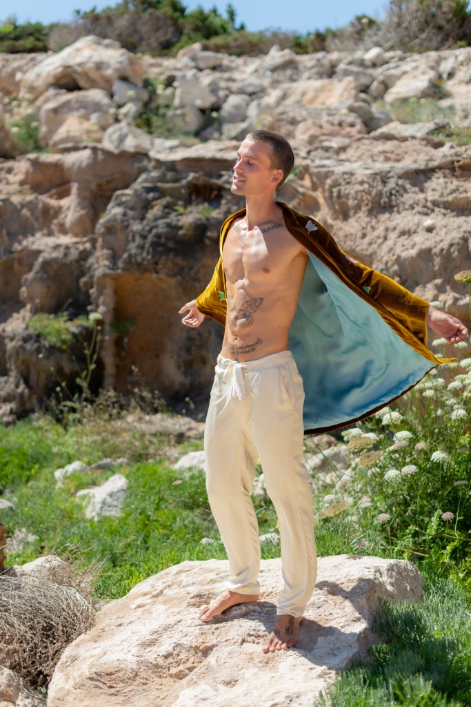 A young man standing barefoot on a rock outdoors, wearing beige trousers and a brown jacket with a blue lining, with a rocky landscape and greenery in the background.