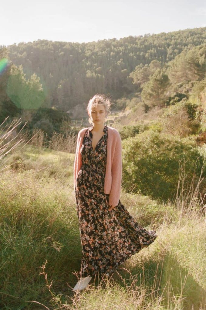 Elegant woman in floral maxi dress and pink cardigan outdoors in nature, featuring lush green trees, mountain backdrop, and sunlight, capturing peaceful summer scenery at La Galeria Elefante.