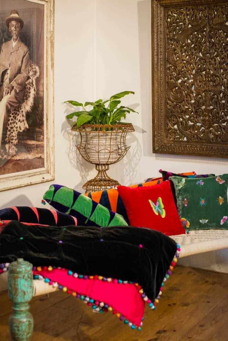Colorful bohemian-style sofa with vibrant pillows and pom-pom trim, vintage artwork, intricate wooden wall decor, and lush green plant at La Galeria Elefante.