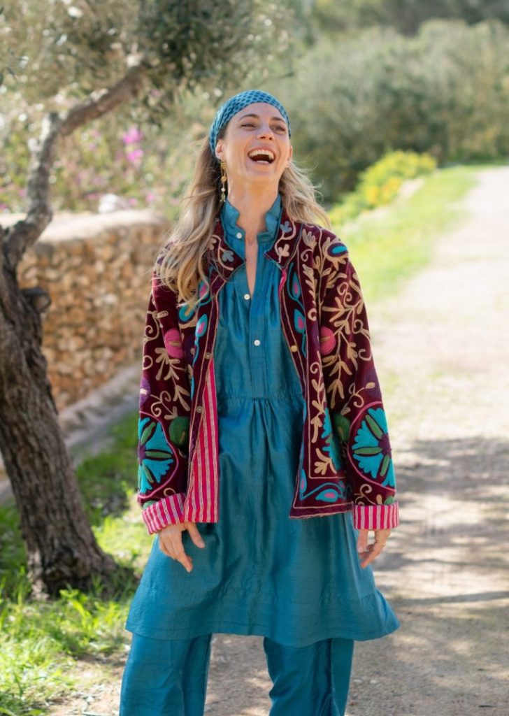 Vibrant woman smiling outdoors in colorful embroidered jacket, blue dress, and headscarf, standing on a sunny nature trail with greenery and trees, promoting La Galeria Elefante.