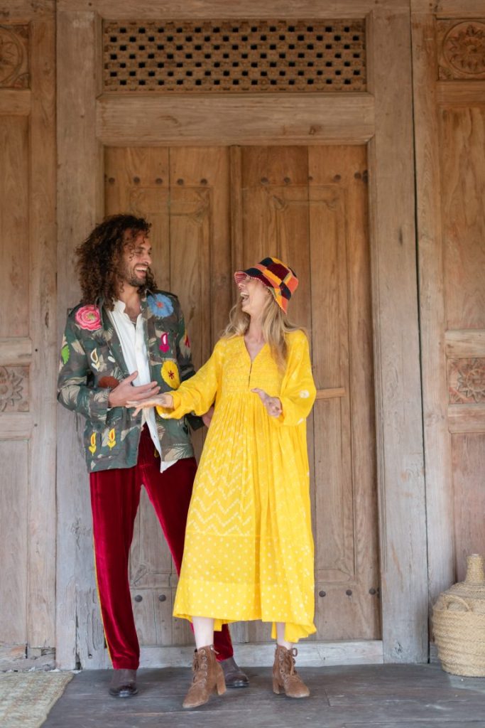 Colorful couple enjoying laugh at La Galeria Elefante, vibrant fashion, joyful atmosphere, unique decor, art-filled space, Bohemian style, artistic ambiance. Woman in a yellow dress with a multi coloured bucket hat with a man with a floral jacket with red velvet trousers