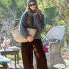 A woman wearing Aladdin Joggers in Velvet, cozy sweater, and beanie, standing outdoors in a lush garden setting with outdoor furniture and vibrant greenery.