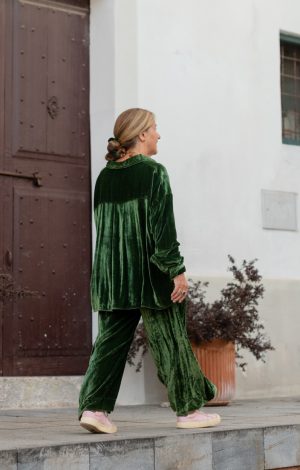 La Galeria Elefante Ibiza Woman in green velvet loungewear walking past a white building with potted plant.