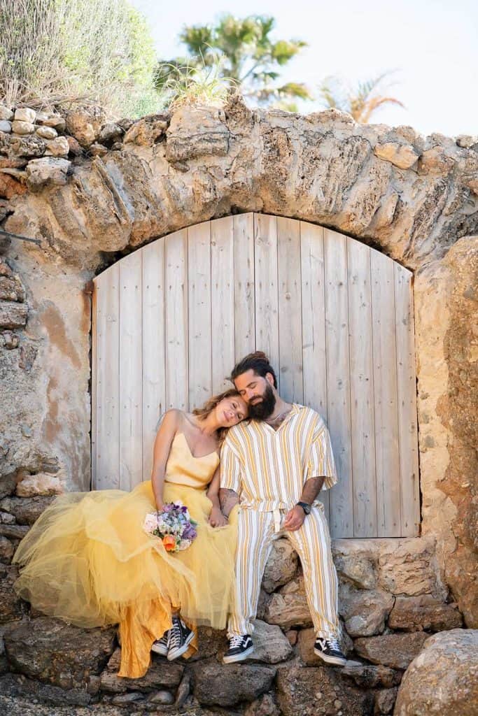 La Galeria Elefante Ibiza Stylish couple in yellow outfits sitting by stone arch in Ibiza, bohemian style, sunny day.