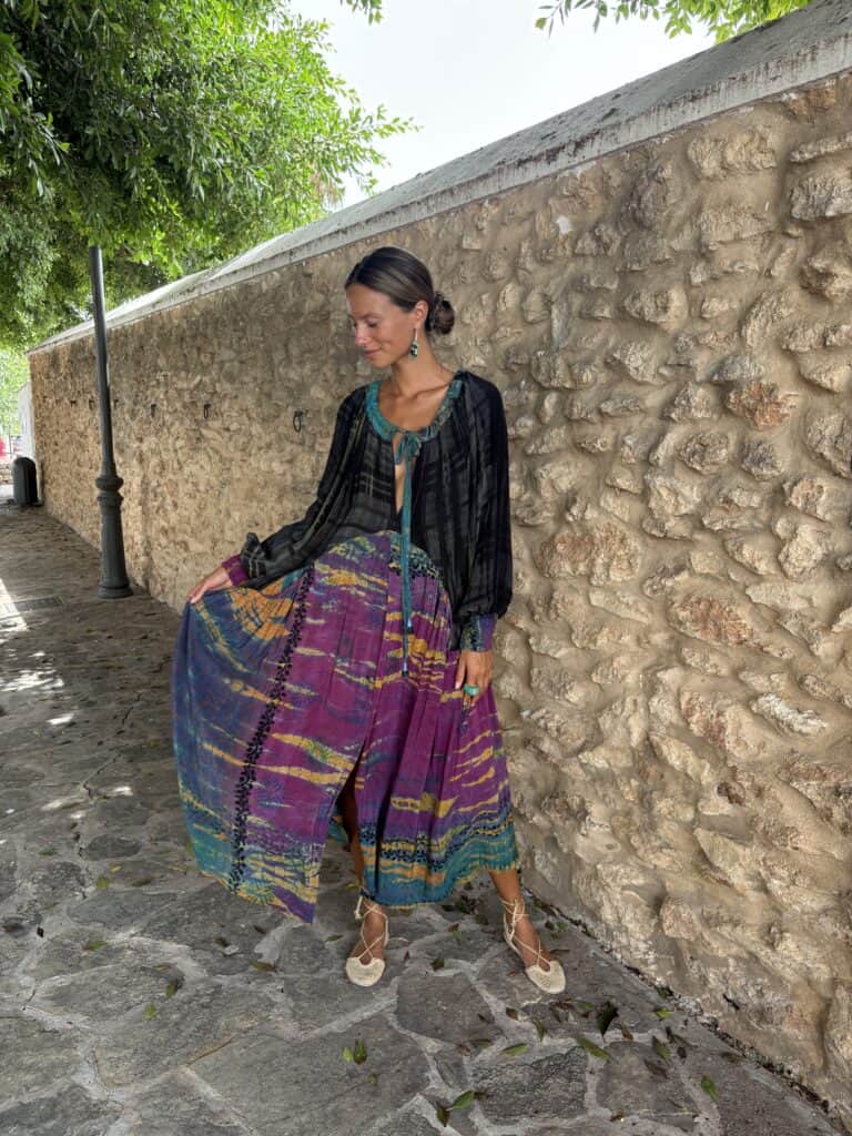 La Galeria Elefante Ibiza Woman in bohemian outfit with vibrant skirt and black top by stone wall.